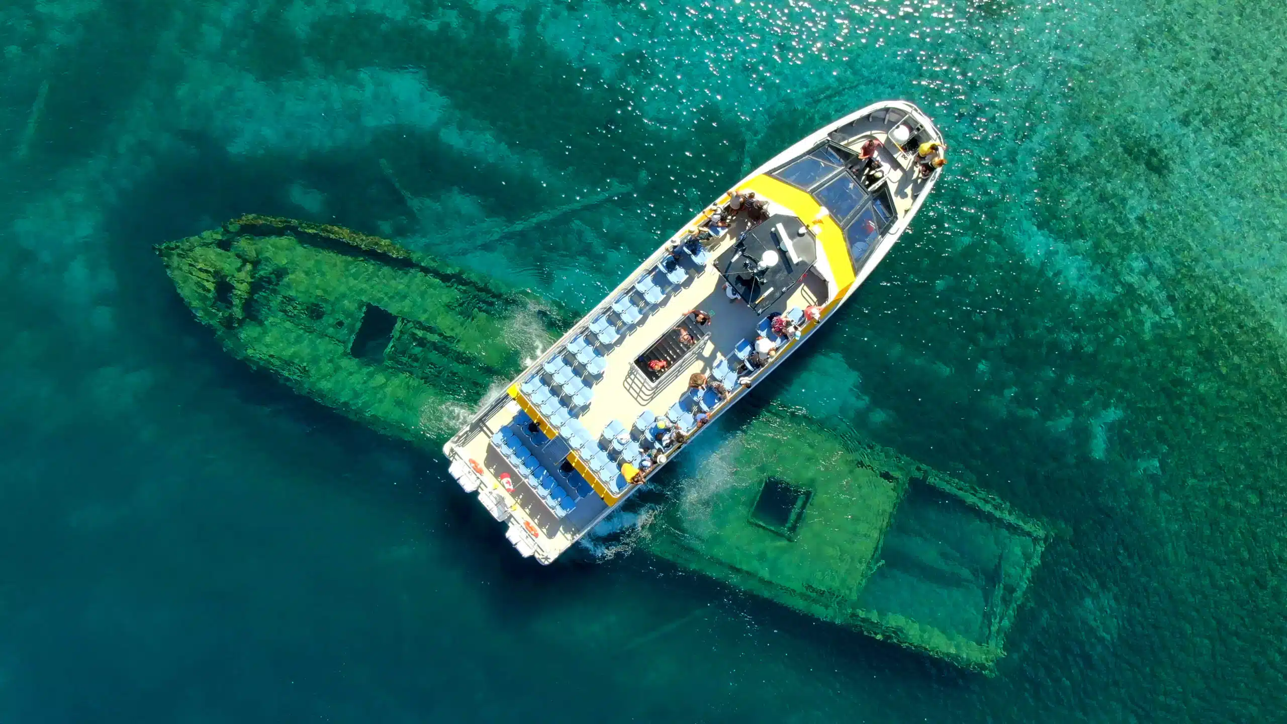 A glass-bottom boat floats above a submerged shipwreck in clear turquoise waters, with passengers observing the marine attraction from above.