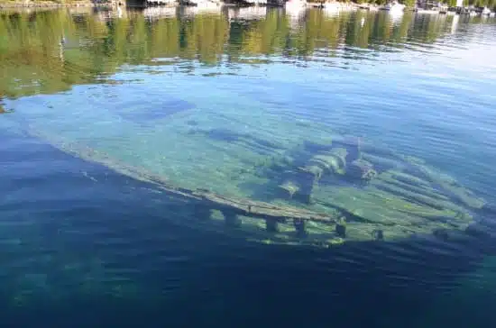 The image shows a submerged shipwreck visible through clear blue water, with trees and reflections on the water's serene surface.