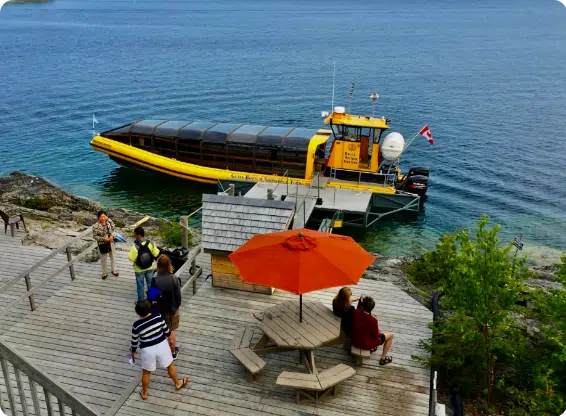 A yellow glass-bottom boat docked near a wooden deck with people, an orange umbrella, and a clear, calm lake background.