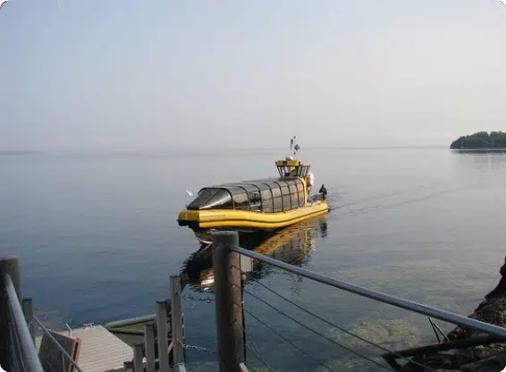 A yellow water taxi approaches a dock on a calm lake, with a person visible onboard. Trees are seen in the distance.