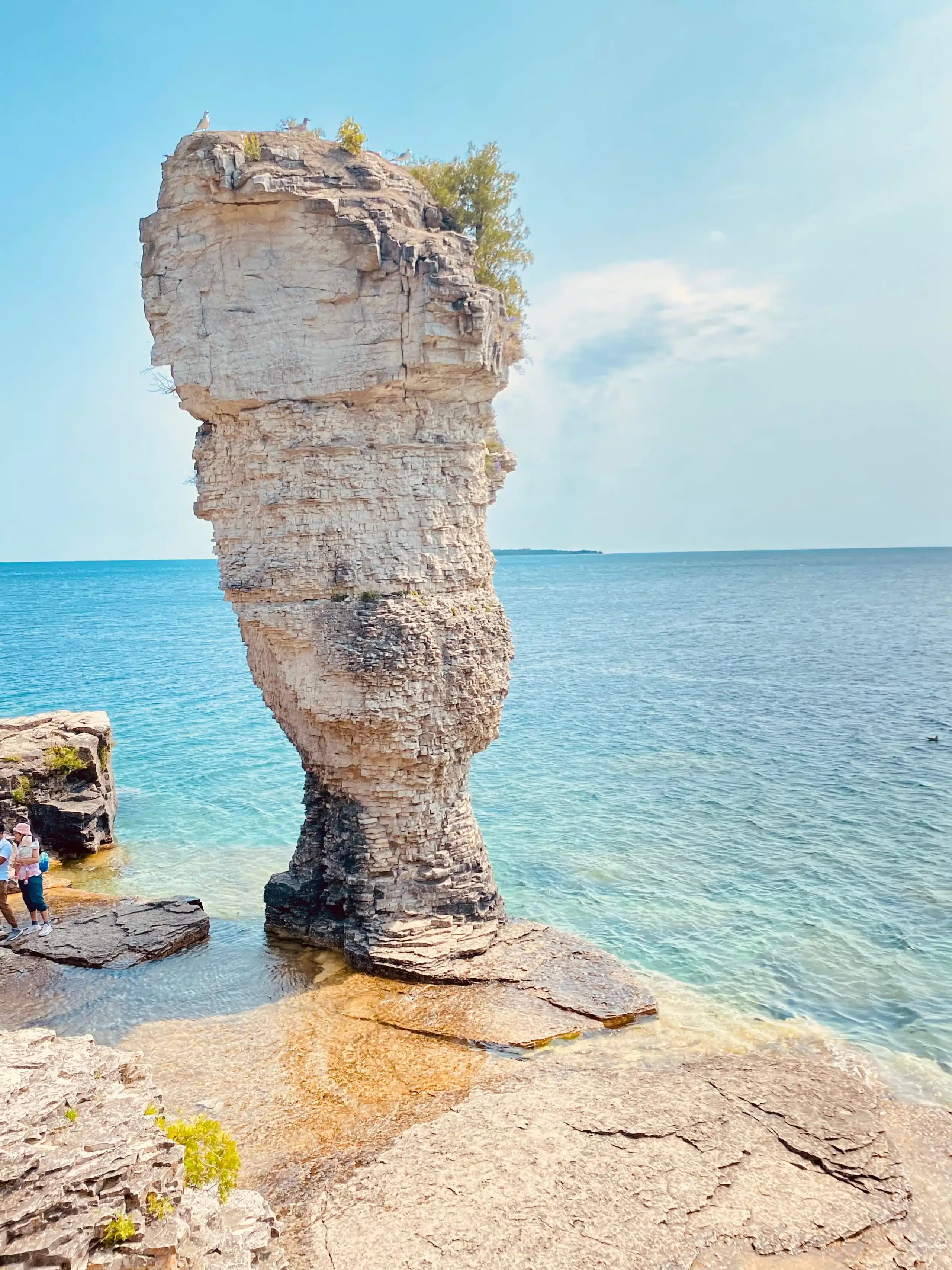 Tall rock formation in shallow seaside water, with two people observing nearby. Clear skies and ocean create a peaceful scene. Flowerpot Island. Flowerpot Rock Formations. Bruce Anchor Cruises. Tobermory. Fathom Five National Marine Park. Bruce Peninsula National Park.