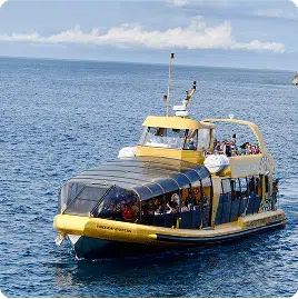 A yellow, glass-roofed ferry on calm blue water, carrying people. Horizon with distant land and clouds visible in the background. No landmarks identified.
