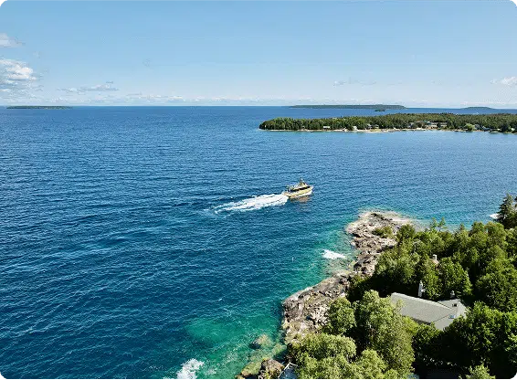 A boat travels across clear blue water near a rocky shoreline and lush greenery, under a clear sky, with distant islands visible.