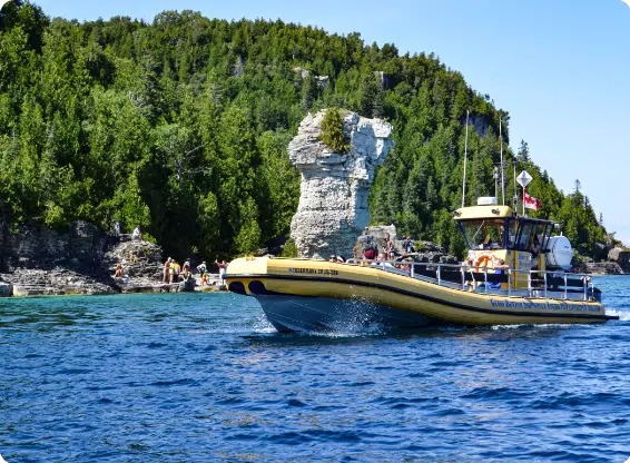 A yellow tour boat navigates clear blue waters near Flowerpot Island, with green cliffs and rock formations in the background. People explore the shoreline.
