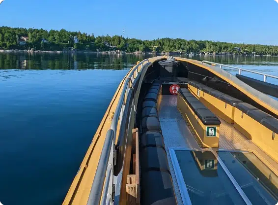 The image shows a small boat with padded seating navigating calm waters surrounded by a lush green shoreline and scattered houses.
