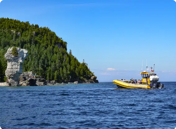 A yellow boat near the Flowerpot Island rock formation in clear water; forested cliff and people visible under a blue sky.