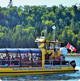 A yellow glass-bottom boat with passengers sails near forested Fathom Five National Marine Park, flying a Canadian flag, under a clear sky.